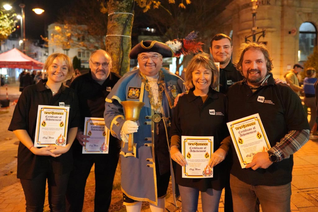 5 ETI team members gather round the Worthing Town Crier, holding their firewalking certificates