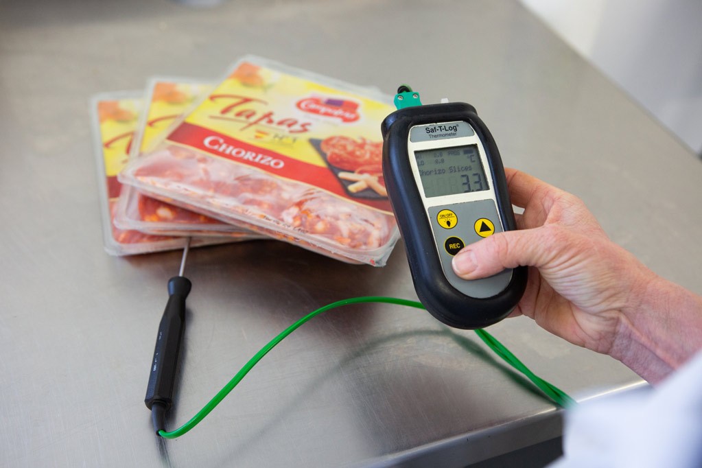 A hand holding a Saf-T-Log thermometer and reading the temperature. The thermometer's wired probe is placed between three packets of chorizo, sitting on a stainless steel countertop.