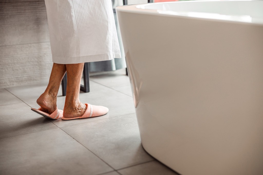 Close up of older woman's legs wearing a white bath robe and pink slippers with the side of the bath in the foreground